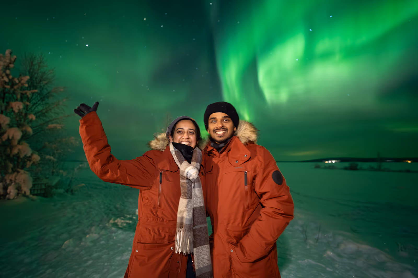 Couple in matching orange jackets beneath swirling green northern lights in March Lapland