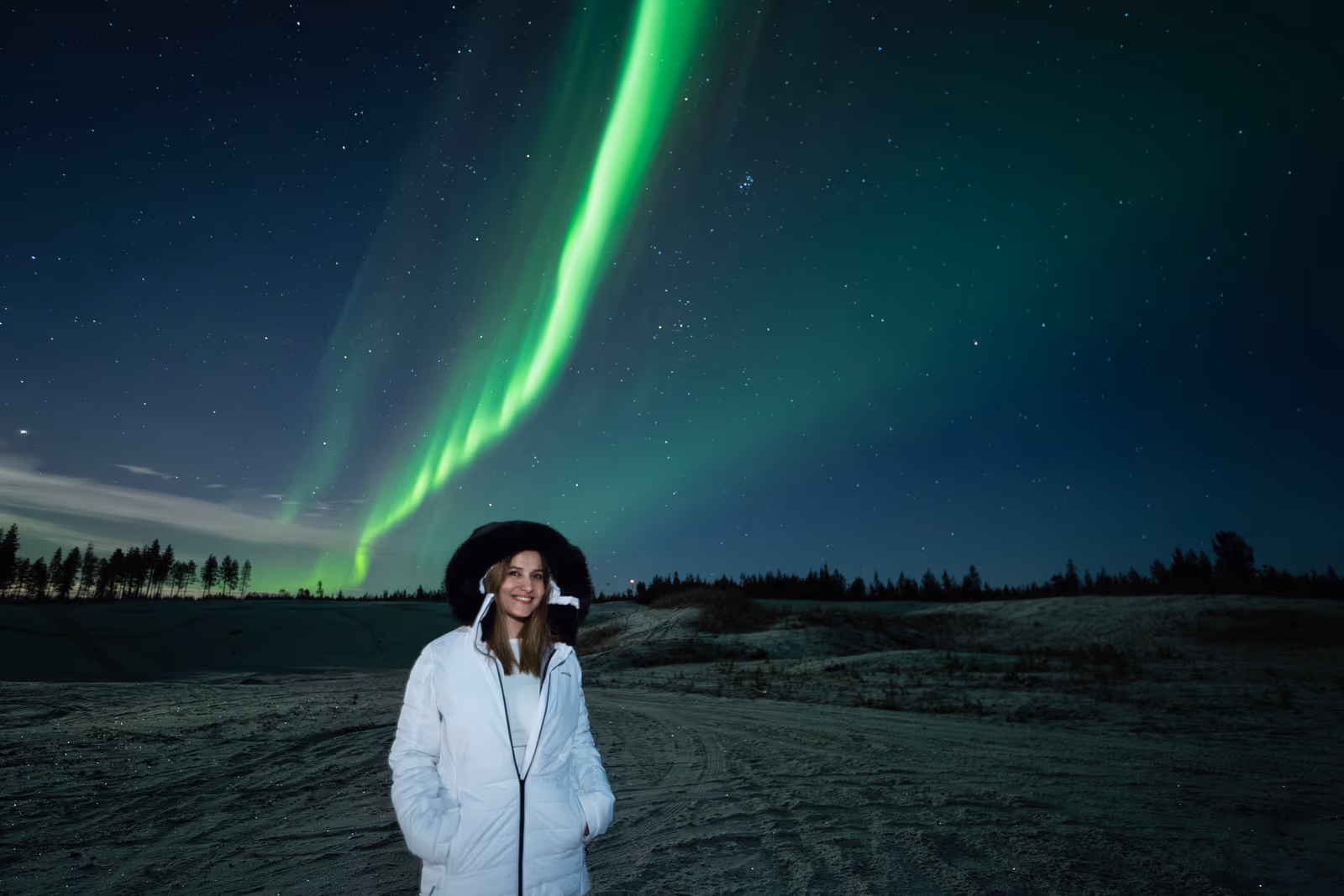 Woman in a white jacket and fur hat beneath a vivid green aurora ribbon in Lapland