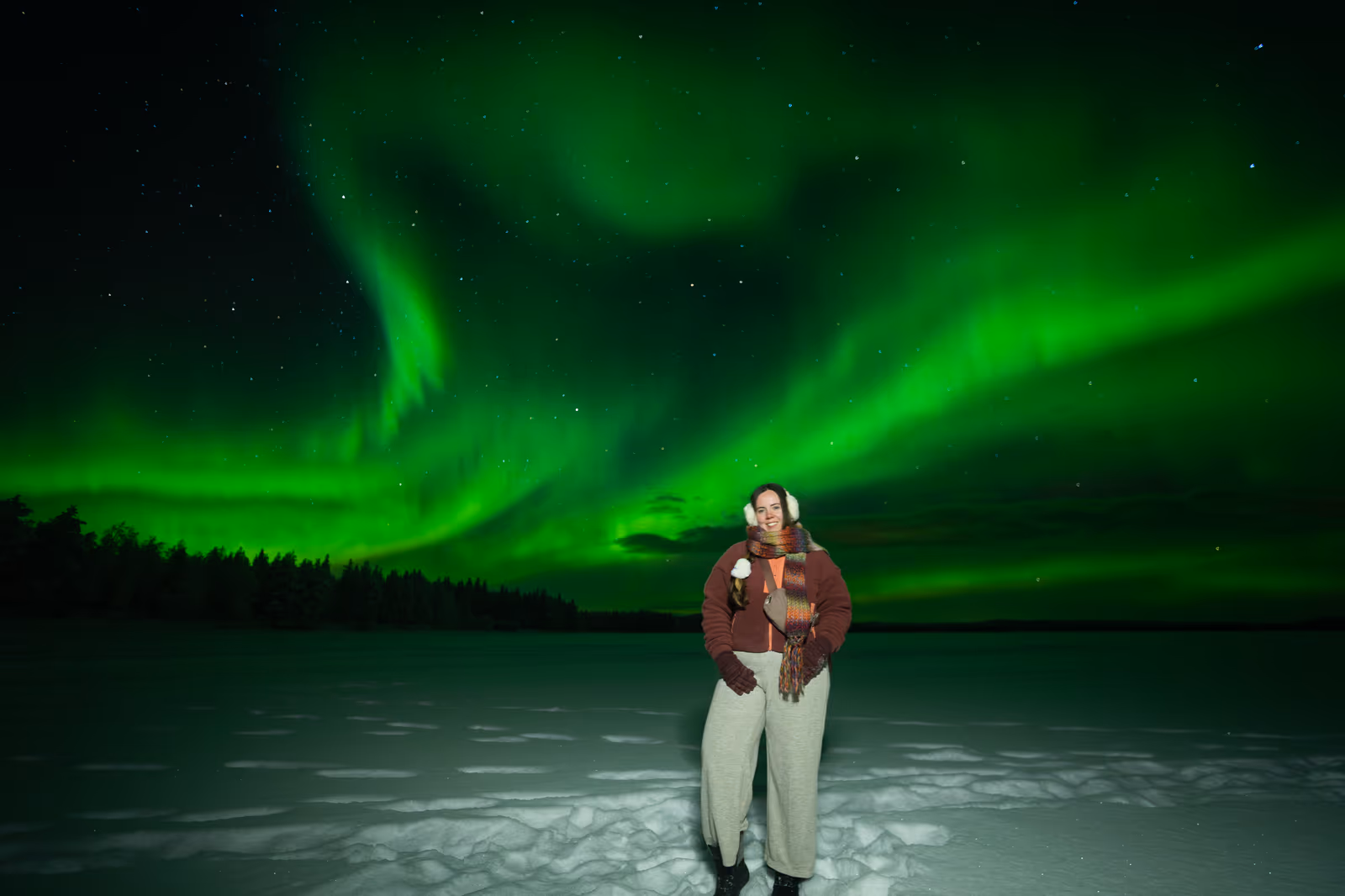 Woman by a frozen lake under a vivid green aurora filling a black, moonless sky